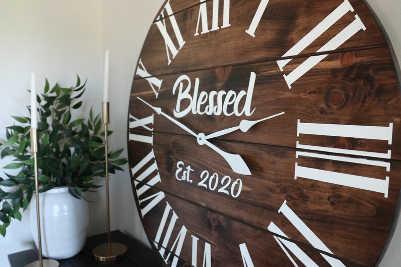 Puede incluir: Un reloj de madera grande y redondo con marcas de n&uacute;meros romanos blancos y manecillas blancas. La esfera del reloj tiene las palabras "Blessed Est 2020" escritas en ella.