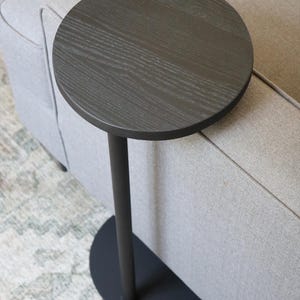 May include: A dark gray, round side table with a single, slender leg and a circular base. The tabletop has a wood grain texture. The table is placed next to a light gray sofa.