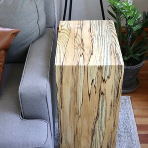 May include: A wooden end table with a natural wood grain finish. The table has a square top and a rectangular base. The table is sitting on a gray rug.