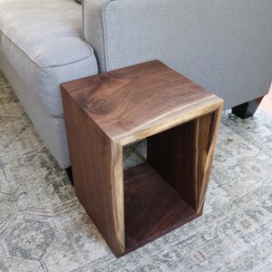 May include: A dark brown wooden end table with a square open shelf. The table has a live edge design and is sitting on a patterned rug.