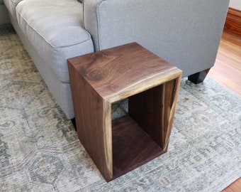 May include: A dark brown wooden end table with a square open shelf. The table has a live edge design and is sitting on a patterned rug.
