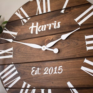 Puede incluir: Un reloj de madera marr&oacute;n con marcas de n&uacute;meros romanos blancos y manecillas blancas. La esfera del reloj tambi&eacute;n incluye el texto "Harris Est. 2015".