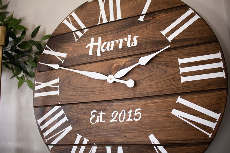 Puede incluir: Un reloj de madera marr&oacute;n con marcas de n&uacute;meros romanos blancos y manecillas blancas. La esfera del reloj tambi&eacute;n incluye el texto "Harris Est. 2015".