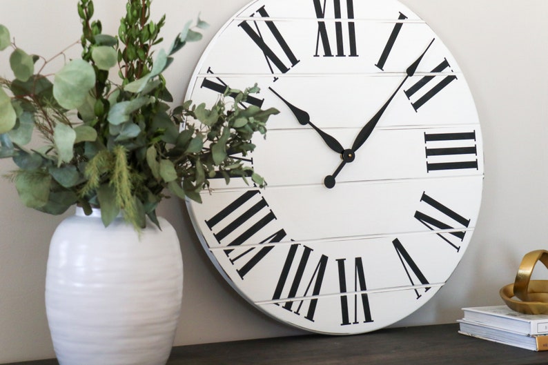 May include: A white round clock with black Roman numeral markings and black hands. The clock is hanging on a wall with a white vase filled with green leafy branches in front of it.