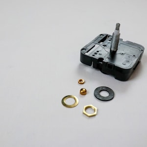 May include: A black clock mechanism with a white shaft and a silver nut. The mechanism is surrounded by several gold-colored metal pieces, including a washer, a nut, and a small round piece.