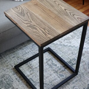 May include: A wooden end table with a black metal frame. The table top has a natural wood grain pattern and is a light brown color. The metal frame is a simple square shape.