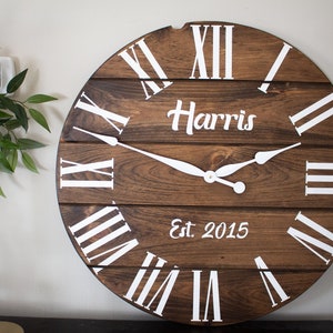 Puede incluir: Un reloj de madera marr&oacute;n con marcas de n&uacute;meros romanos blancos y manecillas blancas. La esfera del reloj tambi&eacute;n incluye el texto "Harris Est. 2015".