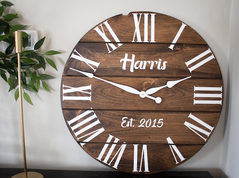 Puede incluir: Un reloj de madera marr&oacute;n con marcas de n&uacute;meros romanos blancos y manecillas blancas. La esfera del reloj tambi&eacute;n incluye el texto "Harris Est. 2015".