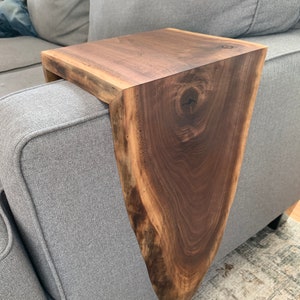 May include: A wooden side table with a live edge design. The table is made of dark brown walnut wood and has a natural, rustic look. It is attached to the arm of a gray couch.