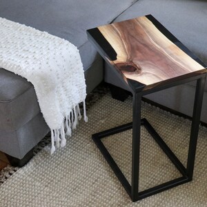 May include: A black metal side table with a wooden top and black epoxy resin inlay. The table has a rectangular top and a square base.