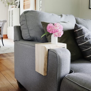 May include: A gray couch with a wooden side table attached to the armrest. A vase with pink flowers sits on the table.