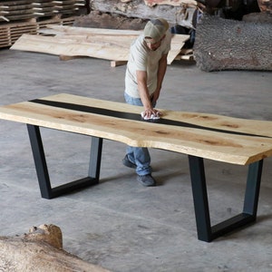 May include: A large, light-colored wooden table with a black resin inlay running down the center. The table has a live edge and sits on black metal legs. A person is wiping the table with a cloth.