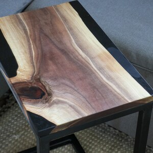 May include: A black metal side table with a wooden top. The top is made of a natural wood with a black epoxy resin inlay. The table is approximately 24 inches wide and 18 inches tall.