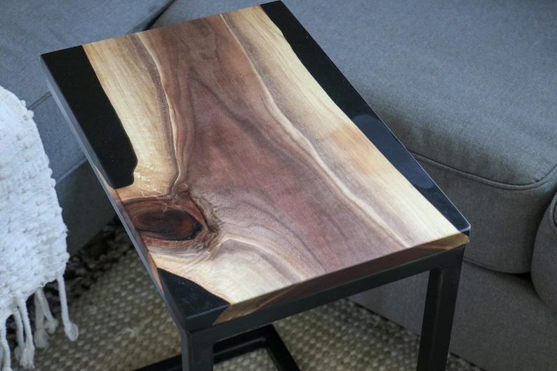 May include: A black metal side table with a wooden top. The top is made of a natural wood with a black epoxy resin inlay. The table is approximately 24 inches wide and 18 inches tall.