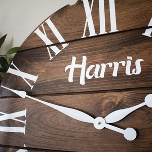 Puede incluir: Una esfera de reloj de madera marr&oacute;n con marcadores de hora en n&uacute;meros romanos blancos y manecillas blancas. La esfera del reloj tiene el nombre "Harris" escrito en letras blancas cursivas.