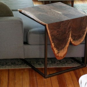 May include: A dark brown wooden side table with a unique, organic shape. The table has a black metal frame and is sitting next to a gray couch.