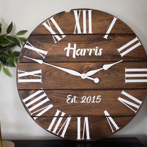Puede incluir: Un reloj grande, redondo y de madera con n&uacute;meros romanos y fondo blanco. La esfera del reloj es marr&oacute;n y tiene un aspecto desgastado. El reloj tiene una inscripci&oacute;n blanca que dice "Harris Est. 2015".
