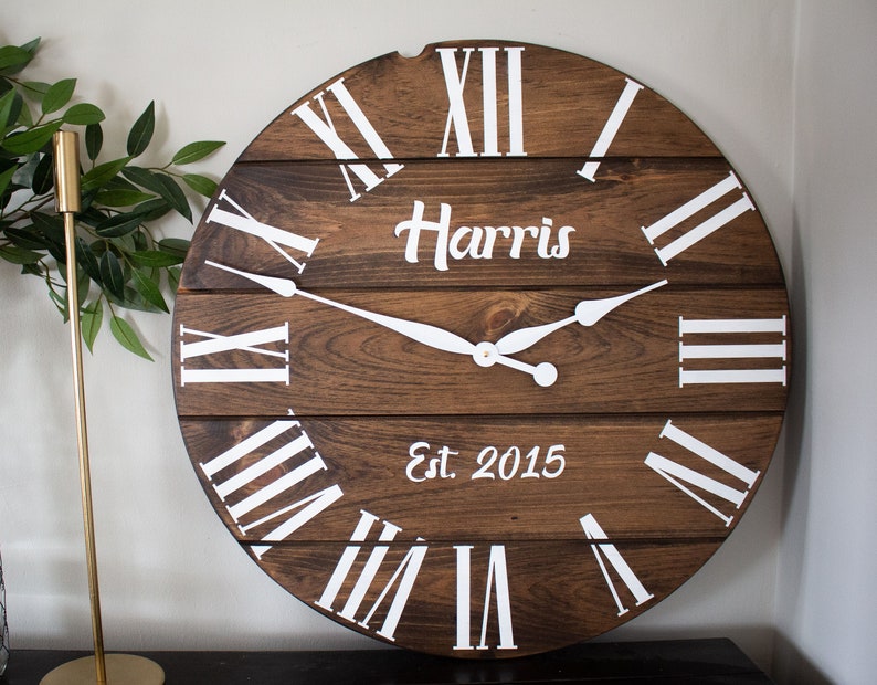 Puede incluir: Un reloj grande, redondo y de madera con n&uacute;meros romanos y fondo blanco. La esfera del reloj es marr&oacute;n y tiene un aspecto desgastado. El reloj tiene una inscripci&oacute;n blanca que dice "Harris Est. 2015".