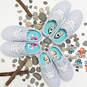 May include: Five pairs of light blue and pink striped canvas shoes with colourful personalised labels inside. The labels feature a variety of designs, including a football, a robot, a monkey, and a purple monster. The shoes are surrounded by small grey stones and two brown sticks.