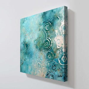May include: Square canvas art with an abstract design. The artwork features a blend of blue and green hues with white circular patterns resembling ripples or raindrops. The overall effect is a textured, watercolor-like appearance.