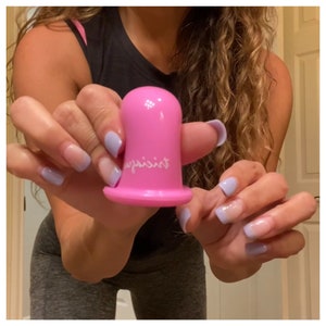 May include: A pink silicone suction cup with the word "against" written on it. The cup is being held by a person with light blue nail polish.