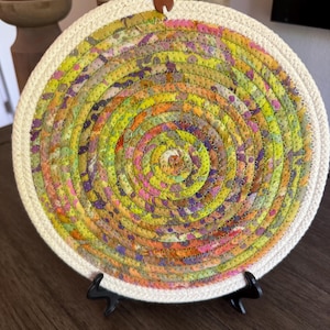May include: A circular trivet made of coiled fabric rope featuring a multicolored pattern of yellow, green, orange, and purple hues. The outer edge is finished with a thick, cream-colored rope border and includes a small brown leather tab loop.