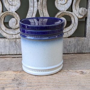 May include: A ceramic jar with a dark blue lid and a white and blue body. The jar has a cylindrical shape with a decorative band near the base. The jar is sitting on a wooden surface.