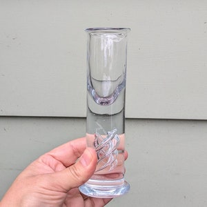 May include: A clear glass shot glass with a swirl design on the side. The glass has a rounded bottom and a straight, cylindrical top.