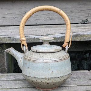 May include: A gray and brown ceramic teapot with a light blue glaze and a bamboo handle. The teapot is sitting on a wooden surface.