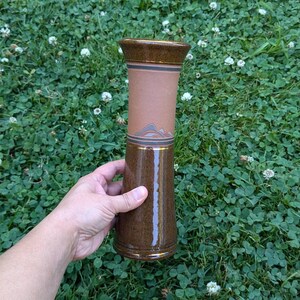May include: A brown ceramic vase with a geometric design and a light brown glaze. The vase is tall and slender, with a rounded base and a flared top.