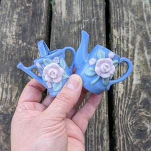 May include: Two small blue ceramic watering cans with pink and green floral designs. The watering cans are held in a hand.