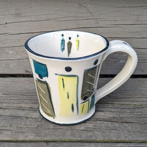 May include: A white ceramic mug with a handle. The mug has a geometric design in shades of yellow, green, brown, and black.