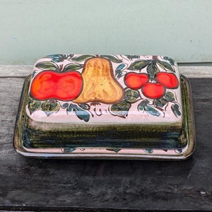 May include: A ceramic butter dish with a lid. The dish is decorated with a pink background and a green, red, and yellow fruit design. The lid has a green rim.