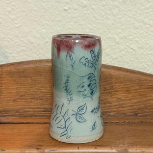 May include: A small, hand-painted ceramic vase with a light blue glaze and a red rim. The vase is decorated with a blue floral pattern.