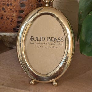 May include: An oval-shaped solid brass picture frame with a polished finish, featuring a small circular hanging loop at the top and two ball-shaped feet at the base. The center insert reads: SOLID BRASS, hand polished & lacquer coated, 2 1/2" x 3 1/2".