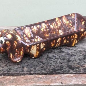 May include: A ceramic dachshund-shaped dish with a brown and cream mottled glaze. The dog has large, expressive eyes, a black nose, and perky ears. The dish is rectangular with a raised rim, perfect for serving snacks or holding small items.