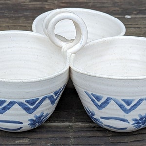 May include: A white ceramic serving dish with three bowls and a handle. The bowls have a blue and white geometric pattern.