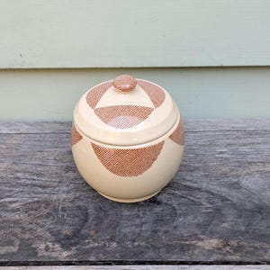 May include: A cream-colored ceramic jar with a lid, featuring a repeating pattern of reddish-brown diagonal stripes within curved shapes. The jar has a rounded body and a small, knob-like handle on the lid. The jar is sitting on a weathered wooden surface.