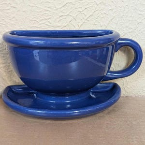 May include: A glossy, deep blue ceramic wall planter shaped like a half-cup and saucer. The piece features a flat back for mounting, a rounded bowl, a curved handle on the right side, and a semi-circular base that mimics the look of a saucer.