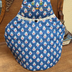 May include: A quilted fabric tea cozy shaped like a cat, featuring a blue background with a repeating pattern of small red and white floral motifs. It has two green button eyes, yellow embroidered whiskers, and a cream lace collar with a black heart.