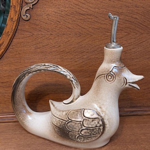 May include: A ceramic duck-shaped oil or vinegar dispenser. The dispenser is primarily beige with brown accents on the wings and handle. It features a silver spout and a decorative design around the neck.