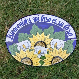 May include: Oval ceramic welcome sign with a blue border and the Spanish text "Bienvenidos mi casa es su casa." The sign features yellow sunflowers, white calla lilies, and green leaves. Two metal loops are attached for hanging.