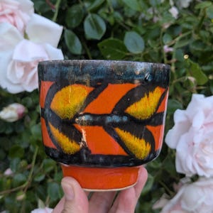 May include: A ceramic cup featuring a vibrant orange base and a wide band decorated with a repeating black and yellow leaf-like pattern. The upper rim is finished with a dark, metallic-toned glaze, and the bottom section is a solid, bright orange.
