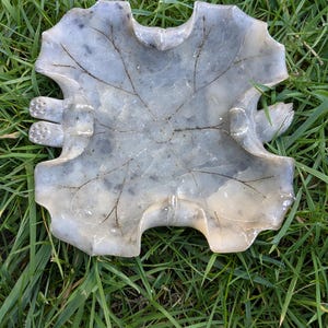 May include: A gray, leaf-shaped decorative dish with a textured surface and visible veins. The dish has a unique, organic design and is resting on a bed of green grass. The dish has a width of approximately 10 inches.