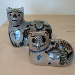 May include: Two ceramic cat figurines. The larger cat is gray with brown and black accents and floral designs. The smaller cat is tan with blue, brown, and green floral patterns. Both have stylized faces.