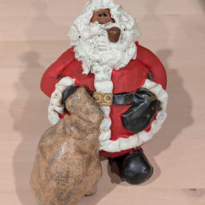 May include: A ceramic Santa Claus figurine in a red suit with white trim, a black belt, and black boots. The figurine has a white beard, a red hat, and is holding a brown sack. A brown pipe is in his mouth.