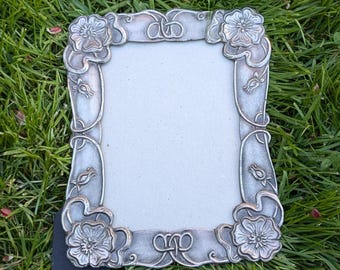 May include: A silver picture frame with a floral design. The frame has a raised design of flowers and leaves around the edges. The frame is sitting on a bed of green grass.