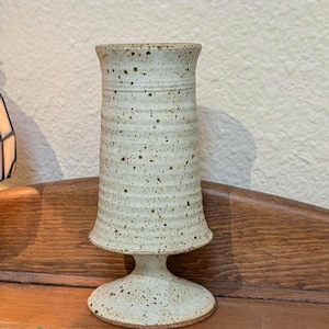 May include: A white ceramic vase with a textured surface and a pedestal base. The vase has a speckled appearance and is a light cream color.