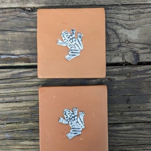 May include: Two terracotta tiles with a white frog design. The frogs are facing the same direction and have a speckled pattern.