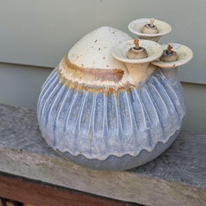 May include: A ceramic oil lamp with a rounded, ribbed body featuring a blue and grey crackle glaze. The top section is cream-colored with brown speckles and includes three small, funnel-shaped openings, each fitted with a central wick.